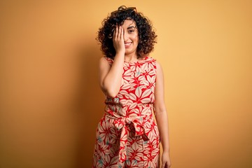 Young beautiful curly arab woman on vacation wearing summer floral dress and sunglasses covering one eye with hand, confident smile on face and surprise emotion.