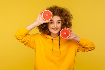 Happy lively energetic woman with fluffy curly hairstyle holding half slice grapefruit and smiling to camera, healthy raw fresh vitamin food concept. indoor studio shot isolated on yellow background © khosrork