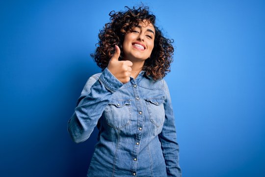 Young beautiful curly arab woman wearing casual denim shirt standing over blue background doing happy thumbs up gesture with hand. Approving expression looking at the camera showing success.
