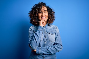 Young beautiful curly arab woman wearing casual denim shirt standing over blue background looking confident at the camera with smile with crossed arms and hand raised on chin. Thinking positive.