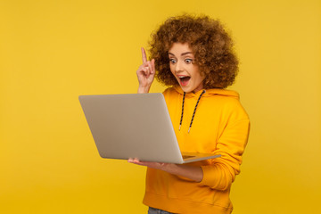 Wondered excited bright woman with curly hair in urban style hoody holding laptop and pointing...