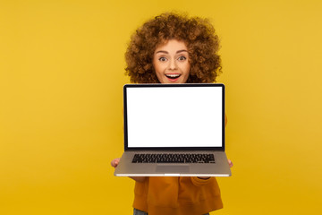 Fototapeta premium Excited amazed happy woman with fluffy curly hair holding laptop with empty screen, showing mock up, blank display for web advertise, commercial text. indoor studio shot isolated on yellow background