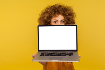 Scared shocked woman with curly hair peeking from behind laptop blank screen with frightened worry...