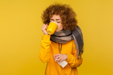 Influenza treatment. Flu-sick woman with curly hair, wrapped in warm scarf, drinking hot tea and holding napkin, taking pills, treating fever with medicine. indoor studio shot, yellow background