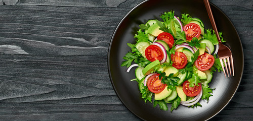 Fresh salad with cherry tomatoes, avocado, cucumeber and lettuce on dish. Healthy vegetarian meal concept. Summer mixed leaves, tomatoes and vegetable salad on black wood, top view.
