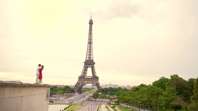 young loving couple standing cuddling together, kissing in the square on the background of the Eiffel Tower. Guy and girl in red dress. Summer. Slow motion. Copy space for text