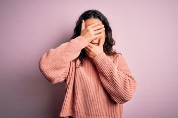 Young beautiful woman with curly hair wearing casual sweater over isolated pink background Covering eyes and mouth with hands, surprised and shocked. Hiding emotion