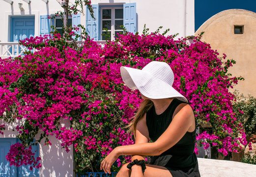 Girl in a white hat and black dress sits in front of a bugambilia plant at the Greek island of Santorini in summer