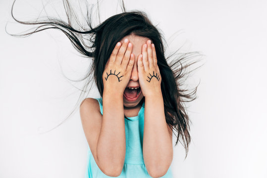 Portrait Of Amazed Little Girl Makes Grimace With Open Mouth And Blowing Hair, Covers Her Eyes With Both Hands With Drawing Doodle Eyes On It. Child Having Fun And Playing Peekaboo. Kid Have Fun.