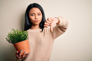 Young beautiful chinese woman holding plant pot standing over isolated white background with angry...