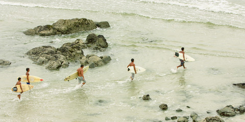 Cinco amigos surfistas carregando pranchas na praia em direção ao mar. Visão aérea.