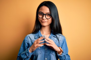 Young beautiful chinese woman wearing casual denim shirt over isolated yellow background Hands together and fingers crossed smiling relaxed and cheerful. Success and optimistic