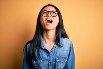 Young beautiful chinese woman wearing casual denim shirt over isolated yellow background angry and mad screaming frustrated and furious, shouting with anger. Rage and aggressive concept.