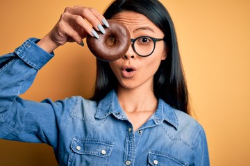 Young beautiful chinese woman holding chocolate donut over isolated yellow background scared in shock with a surprise face, afraid and excited with fear expression