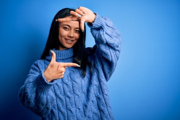 Young beautiful chinese woman wearing casual sweater over isolated blue background smiling making frame with hands and fingers with happy face. Creativity and photography concept.