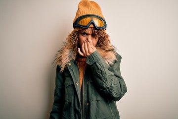 Young african american skier woman with curly hair wearing snow sportswear and ski goggles smelling something stinky and disgusting, intolerable smell, holding breath with fingers on nose. Bad smell