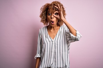 Beautiful african american woman with curly hair wearing striped t-shirt over pink background doing ok gesture shocked with surprised face, eye looking through fingers. Unbelieving expression.