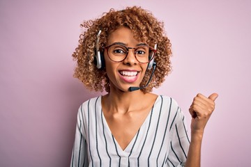 African american curly call center agent woman working using headset over pink background smiling with happy face looking and pointing to the side with thumb up.