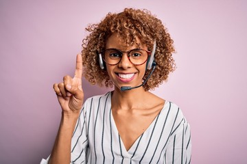 African american curly call center agent woman working using headset over pink background showing...