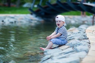 the baby in a white cap sitting by the pond touches cold water with his leg and admires