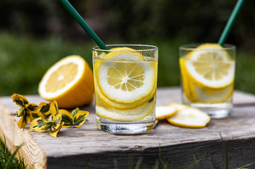Summer drink, glasses of water filled with slices of lemons, stand on a wooden board on the lawn in sunny warm day, the background is blurred, shallow depth of field, selective focus.