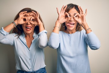 Middle age beautiful couple of sisters standing over isolated white background doing ok gesture like binoculars sticking tongue out, eyes looking through fingers. Crazy expression.