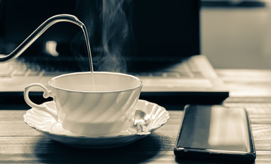 A white cup of coffee with hot smoke in stainless pot, Morning coffee time with notebook computer and work equipment, Normal practice by working at home during covid-19 outbreak around the world.