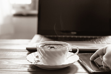 A hand using smartphone with a white coffee cup on the desk in the morning at home,Notebook computer and work equipment, Normal practice by working from home during covid-19 outbreak around the world.