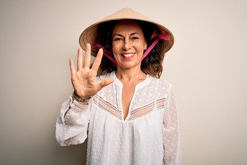 Middle age brunette woman wearing asian traditional conical hat over white background showing and pointing up with fingers number five while smiling confident and happy.