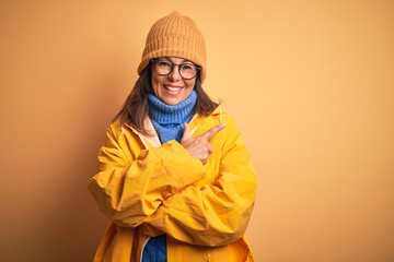 Middle age woman wearing yellow raincoat and winter hat over isolated background cheerful with a smile of face pointing with hand and finger up to the side with happy and natural expression on face