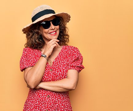 Middle age beautiful woman wearing casual dress and sunglasses over yellow background smiling looking confident at the camera with crossed arms and hand on chin. Thinking positive.