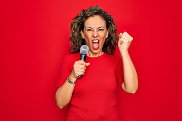 Middle age senior singer woman singing using music microphone over red background annoyed and frustrated shouting with anger, crazy and yelling with raised hand, anger concept