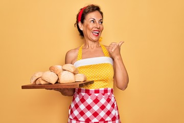 Middle age senior pin up housewife woman wearing 50s style retro dress and apron cooking bread pointing and showing with thumb up to the side with happy face smiling