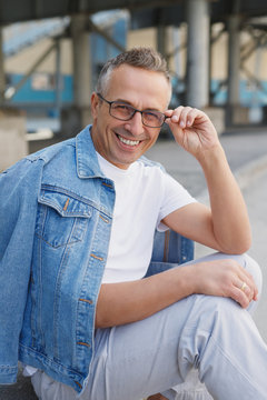 adult man in a white T-shirt, denim jacket and glasses posing against the backdrop of the city during the day
