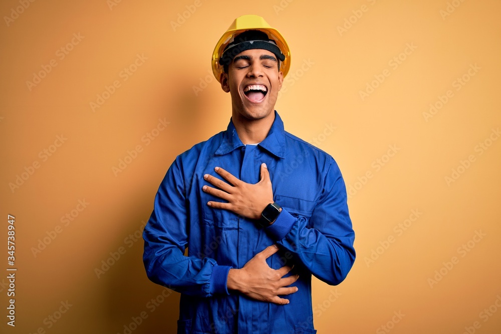 Wall mural young handsome african american worker man wearing blue uniform and security helmet smiling and laug