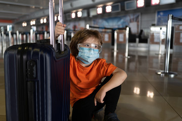 Tired traveler child in protective mask and glasses during coronavirus. Boy stuck with baggage at airport, flight canceled. Children get home during a pandemic and quarantine. Covid 19 concept.