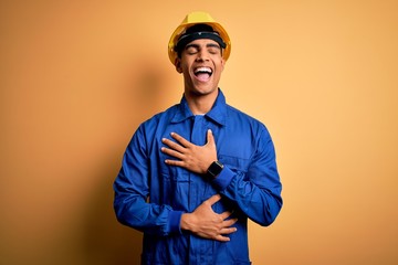 Young handsome african american worker man wearing blue uniform and security helmet smiling and laughing hard out loud because funny crazy joke with hands on body.