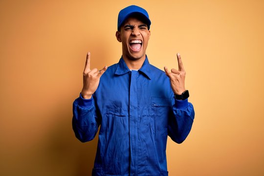 Young african american mechanic man wearing blue uniform and cap over yellow background shouting with crazy expression doing rock symbol with hands up. Music star. Heavy concept.