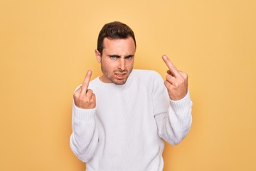 Young handsome man with blue eyes wearing casual sweater standing over yellow background Showing middle finger doing fuck you bad expression, provocation and rude attitude. Screaming excited