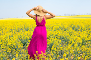 female in hat and magenta dress is standing in the yellow field and admires of the view. view from the back