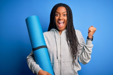 Young african american fitness woman holding yoga mat over isolated blue background screaming proud...