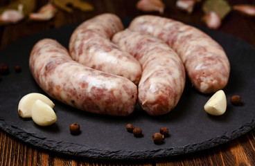 Raw homemade sausages with garlic cloves and spices on stone board on wooden table.