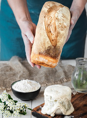 Fototapeta premium Female hands hold freshly baked bread, sourdough and ingredients for baking bread on a white wooden table close-up. hobby, baking wheat bread at sourdough at home. Healthy traditional craft bread.