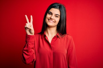 Young beautiful brunette woman wearing casual shirt standing over red background showing and pointing up with fingers number two while smiling confident and happy.