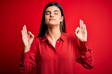 Young beautiful brunette woman wearing casual shirt standing over red background relax and smiling with eyes closed doing meditation gesture with fingers. Yoga concept.