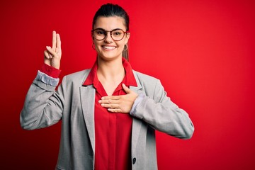 Young beautiful brunette businesswoman wearing jacket and glasses over red background smiling...