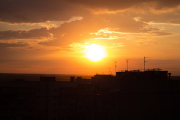 Sunset on the roof of a multi-storey building overlooking a yellow-orange sky in the clouds and the sun, antennas on the roofs.