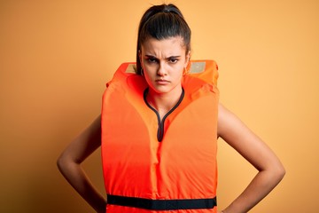 Young beautiful brunette woman wearing orange safe lifejacket over yellow background skeptic and...