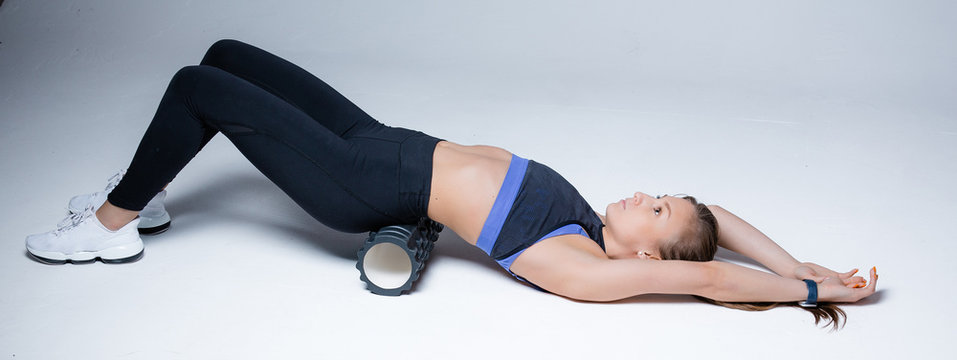The girl massages the fascia of the muscles of the lower back, arms extended up and lying on her back, with a massage roller