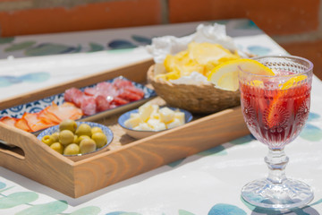 tray filled with tapas with olives, cheese, smoked sausage, slices of ham and a bowl of crisps next to a drinking glass filled with red wine and soda with ice cubes and lemon slices. Sangria concept.